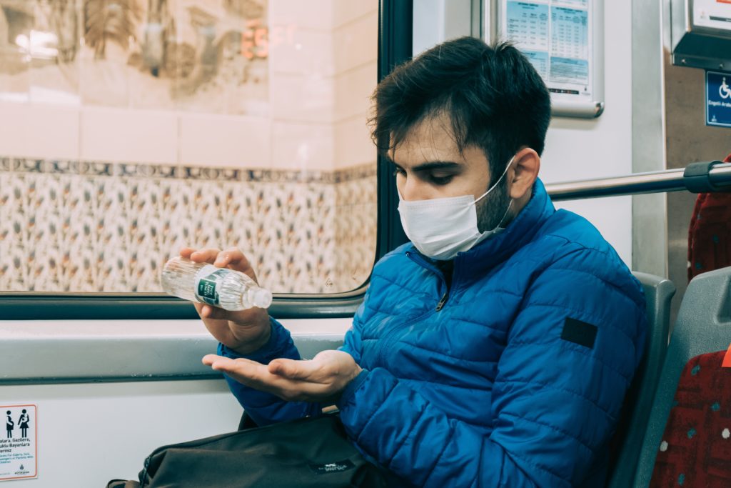 A man sits on a bus using a mask and putting hand sanitizer on his hands. 