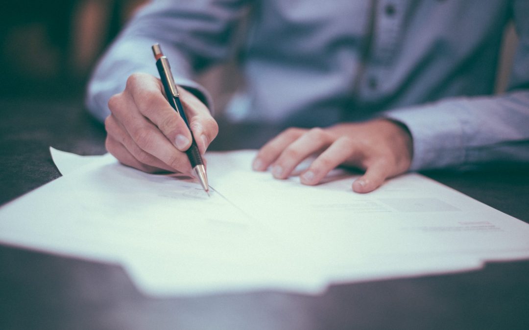 A man wearing a blue button down shirt signs documents on a wooden surface